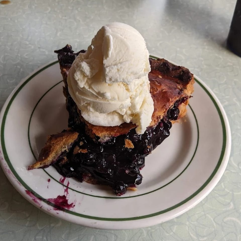A slice of blueberry pie with a large scoop of vanilla ice cream on a white plate.