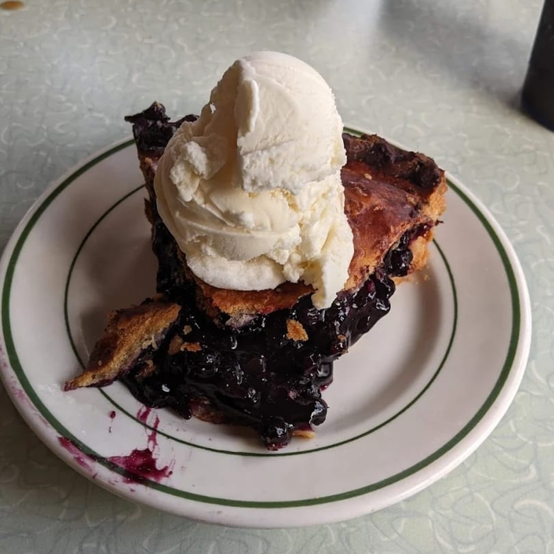 A slice of blueberry pie with a large scoop of vanilla ice cream on a white plate.