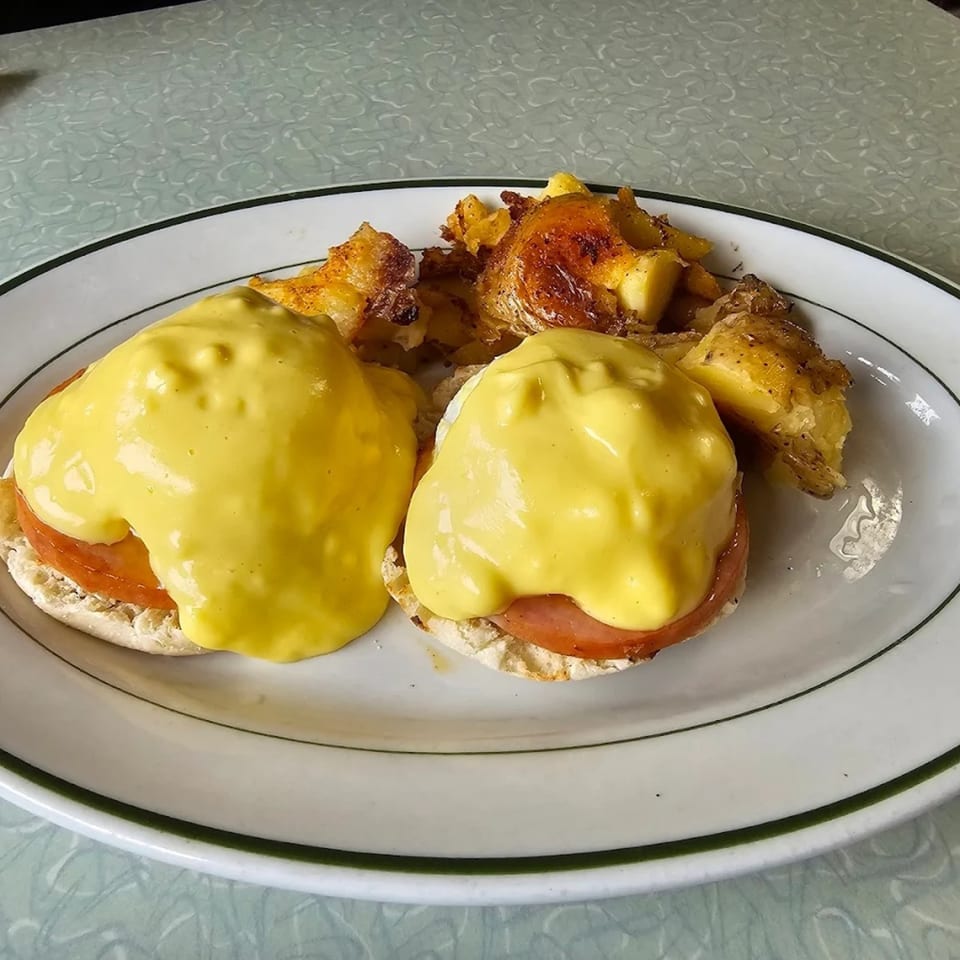 Two eggs benedict with hollandaise sauce and a side of home fries on a white plate.