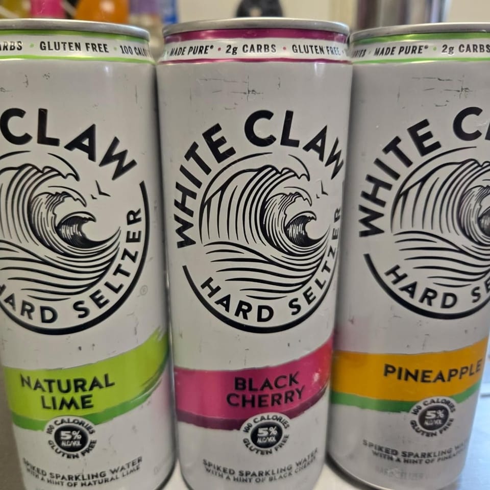 Three White Claw hard seltzer cans in Natural Lime, Black Cherry, and Pineapple flavors.