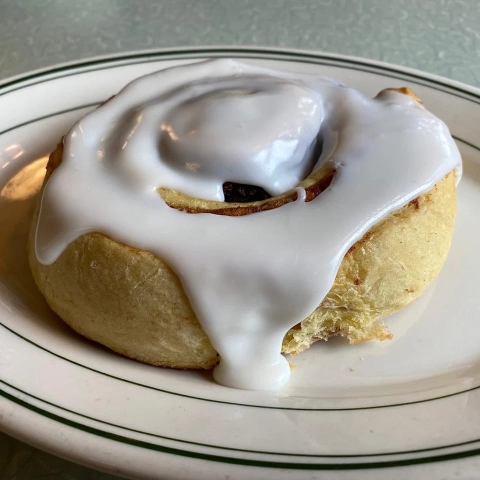 A Chef Darrell’s Mountain Diner cinnamon roll with white icing on a white plate with green stripes.