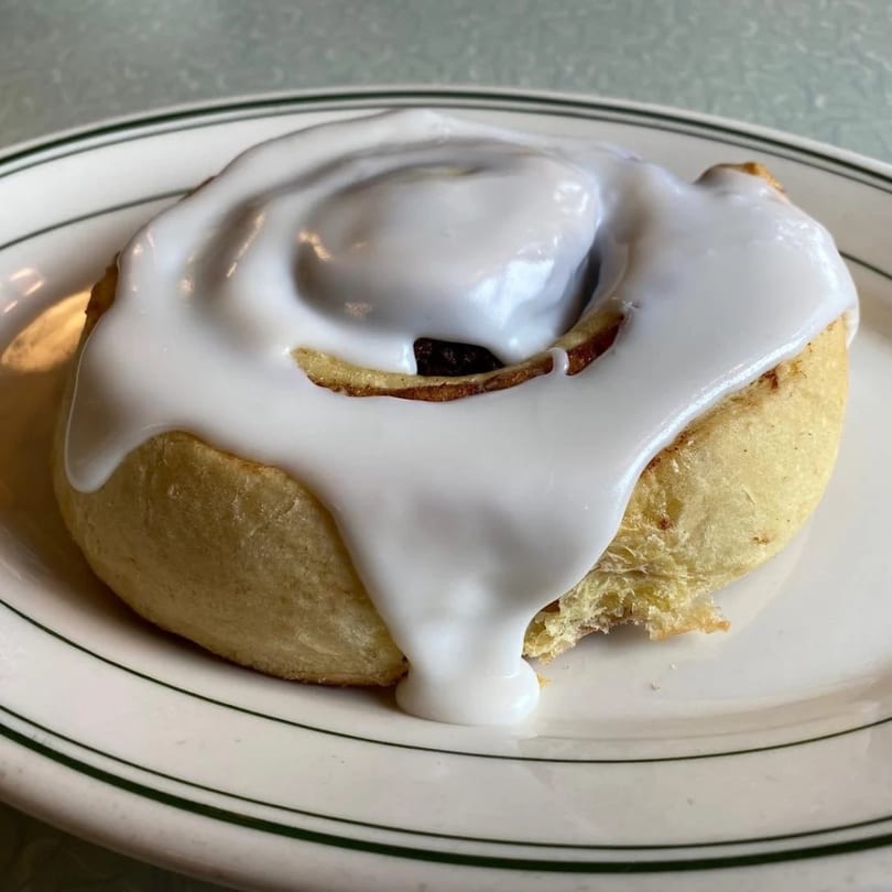 A Chef Darrell’s Mountain Diner cinnamon roll with white icing on a white plate with green stripes.