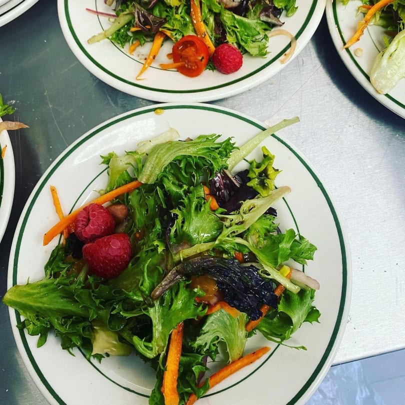 Multiple plates of fresh green salad with raspberries, carrots, and tomatoes on a metal surface.
