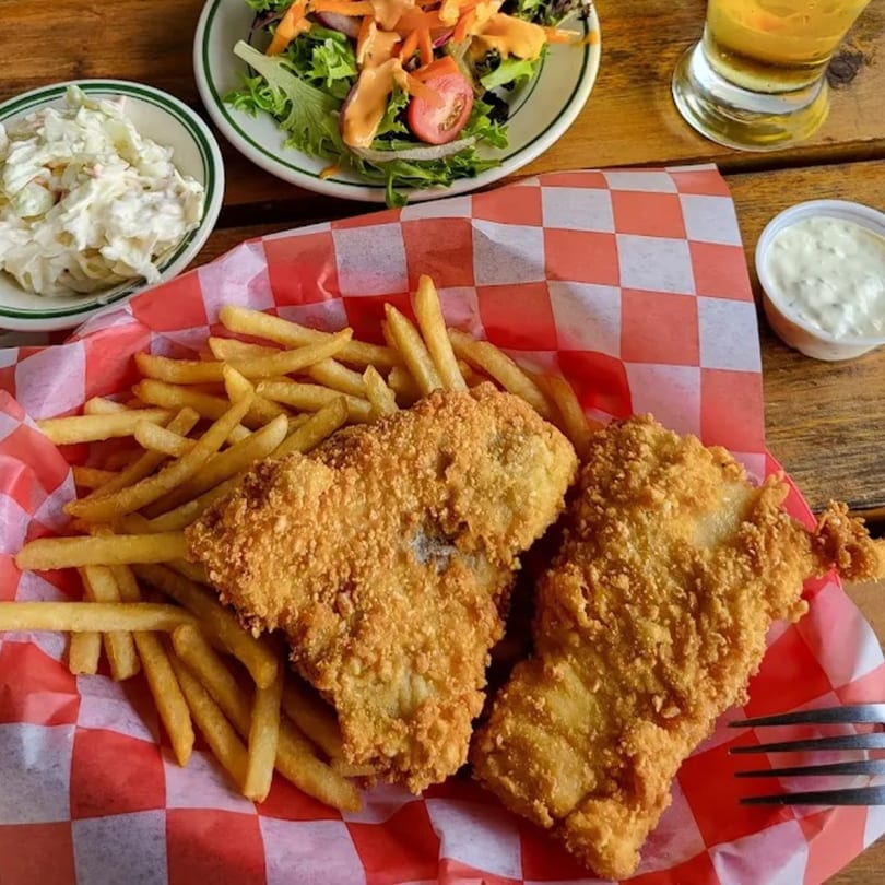 Fish and chips in a red checkered basket, with coleslaw, salad, and a drink.