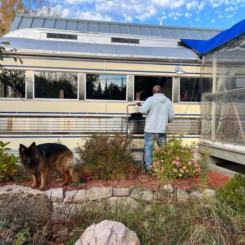 A man and a German Shepherd dog stand outside Chef Darrell's diner, a yellow and silver train car.