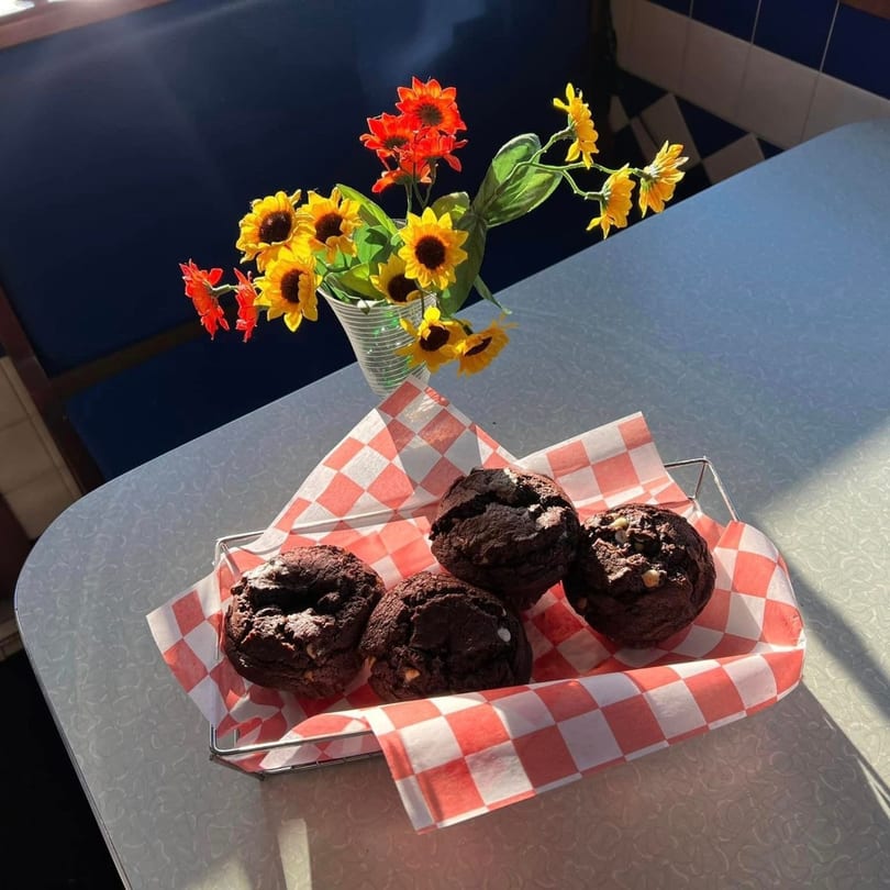 Four chocolate muffins with white chips in a checkered basket on a diner table with sunflowers.