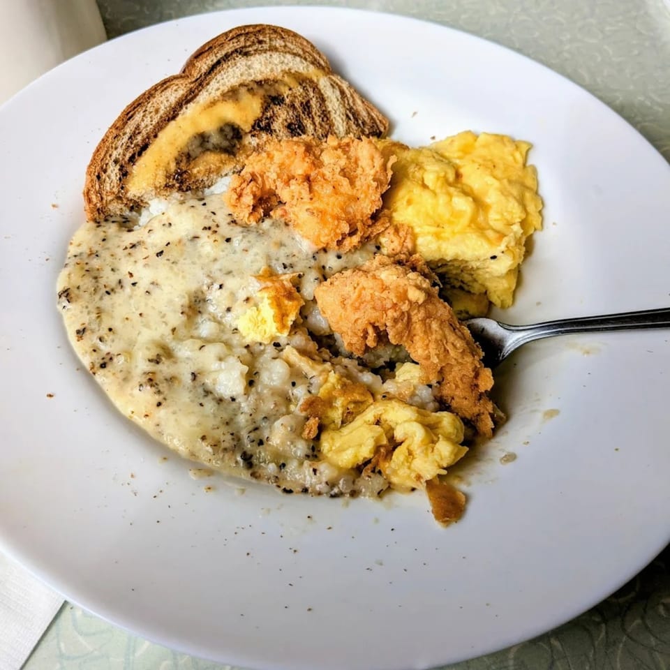 A diner breakfast plate with grits, scrambled eggs, fried chicken, and a slice of toast.