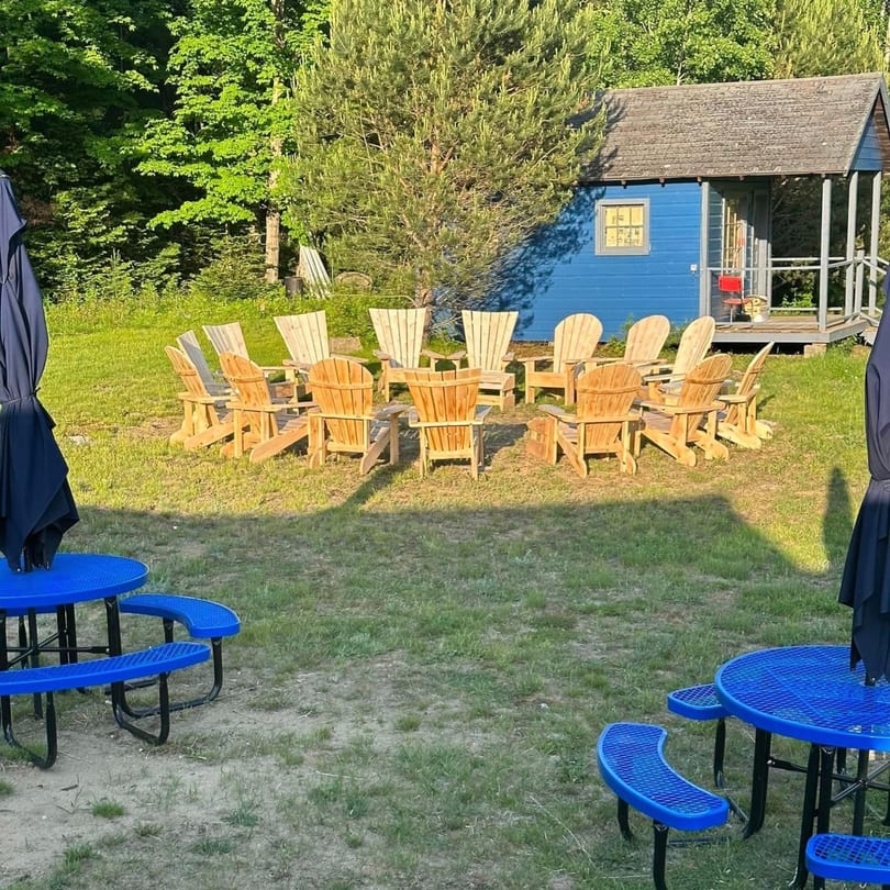 Outdoor seating area with Adirondack chairs in a circle and blue picnic tables near a small blue cabin.
